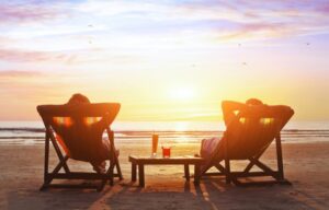 A wealthy retired couple that has been building wealth in their 70s is now relaxing in recliners on a beach at sunset.